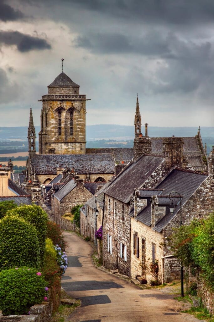 Dramatic stone village street with granite church tower under stormy sky Locronan Brittany France